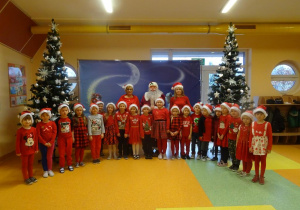 Grupa dzieci stoi z panią dyrektor Marią Królikowską i panią Ewą Janicką, w tle dekoracja świąteczna, choinki.